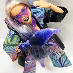May include: A doll with lavender hair and purple-tinted sunglasses, wearing a black jacket with a colorful patterned lining. The doll has a purple lobster claw hand and black lipstick. The outfit has a vibrant, artistic design.