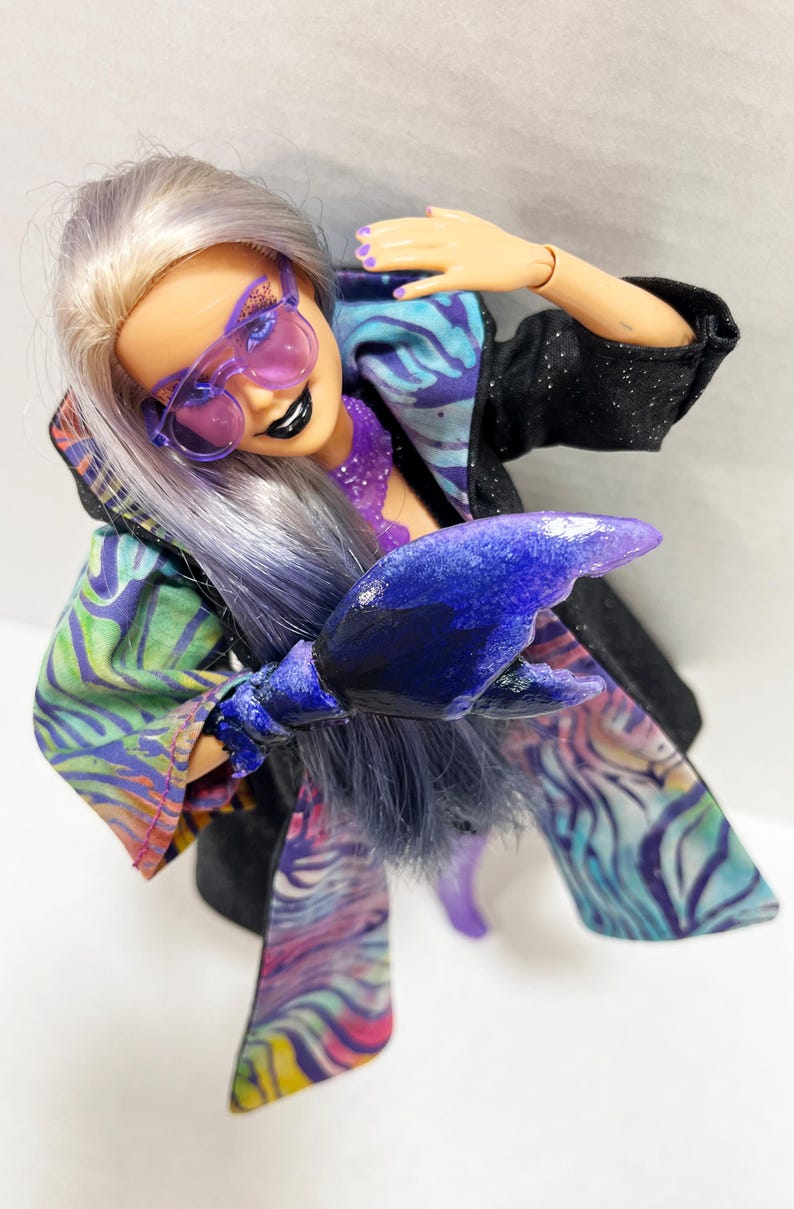 May include: A doll with lavender hair and purple-tinted sunglasses, wearing a black jacket with a colorful patterned lining. The doll has a purple lobster claw hand and black lipstick. The outfit has a vibrant, artistic design.