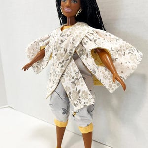 以下が含まれることがあります： 花柄の着物風トップスと、蜂のアクセントが付いたグレーのカプリパンツを着た人形。人形は黒髪で、黄色のサンダルを履いています。この衣装は黄色のトリムが特徴です。