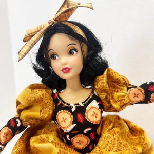 Op de afbeelding: Een pop met zwart haar in een boblijn, met een beige geruite haarstrik. De pop draagt een zwart lijfje met een taartjesprint en een gele rok met een bladpatroon. De mouwen zijn pofmouwen en geel met boorden met een taartjesprint.