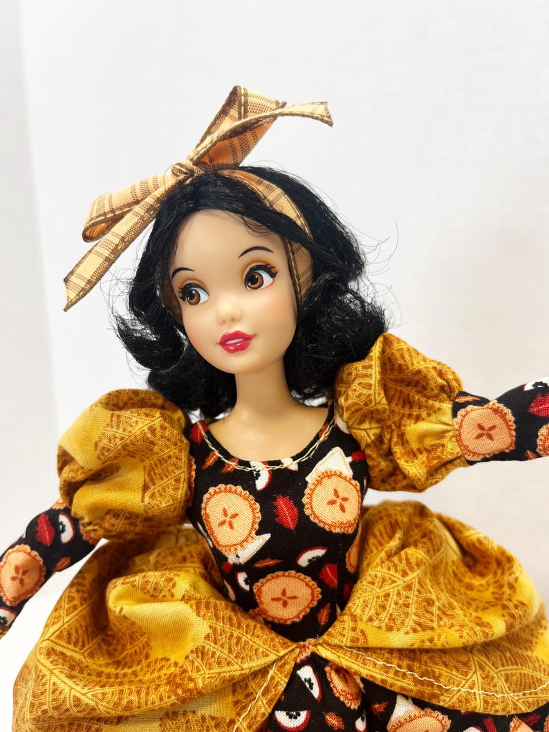 Op de afbeelding: Een pop met zwart haar in een boblijn, met een beige geruite haarstrik. De pop draagt een zwart lijfje met een taartjesprint en een gele rok met een bladpatroon. De mouwen zijn pofmouwen en geel met boorden met een taartjesprint.
