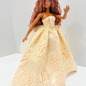 Puede incluir: Una mu&ntilde;eca con pelo largo color casta&ntilde;o rojizo y una corona amarilla. La mu&ntilde;eca lleva un vestido sin tirantes color crema con un estampado de conchas marinas. El brazo de la mu&ntilde;eca est&aacute; levantado en un gesto de saludo. Una enagua de tul a&ntilde;ade volumen al vestido.