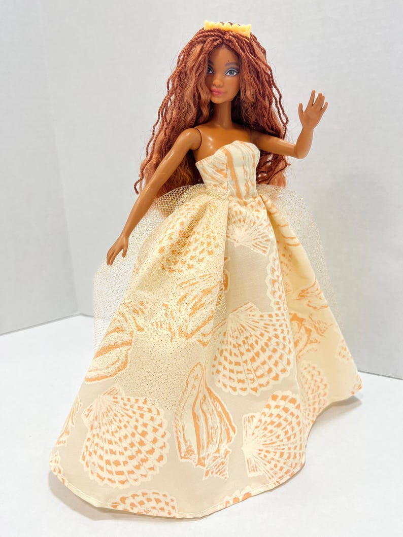 Puede incluir: Una mu&ntilde;eca con pelo largo color casta&ntilde;o rojizo y una corona amarilla. La mu&ntilde;eca lleva un vestido sin tirantes color crema con un estampado de conchas marinas. El brazo de la mu&ntilde;eca est&aacute; levantado en un gesto de saludo. Una enagua de tul a&ntilde;ade volumen al vestido.