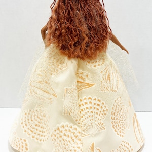 Puede incluir: Una mu&ntilde;eca con cabello largo color casta&ntilde;o rojizo y un vestido color crema adornado con patrones de conchas marinas. El vestido tiene una capa de tul. Una concha marina est&aacute; en la superficie cerca de la mu&ntilde;eca.