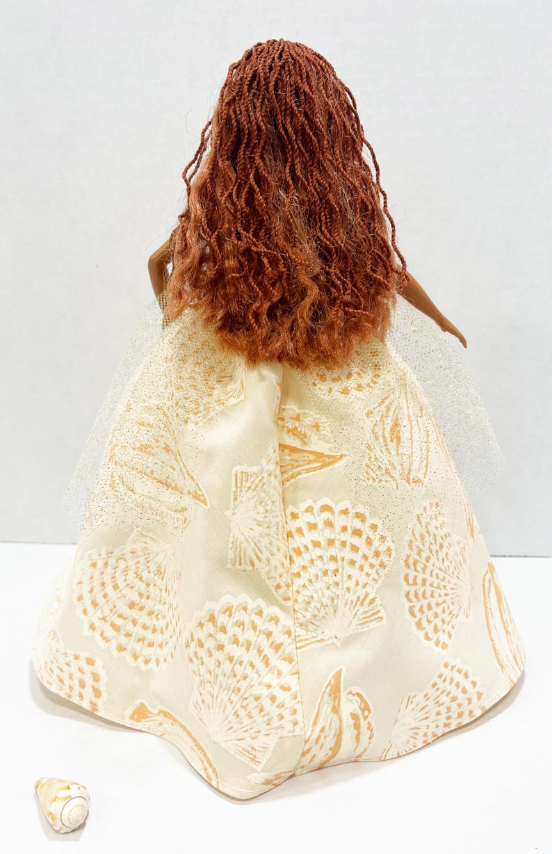 Puede incluir: Una mu&ntilde;eca con cabello largo color casta&ntilde;o rojizo y un vestido color crema adornado con patrones de conchas marinas. El vestido tiene una capa de tul. Una concha marina est&aacute; en la superficie cerca de la mu&ntilde;eca.