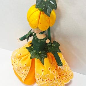 May include: A doll with a yellow pumpkin-shaped head topped with green leaves, wearing a dark green bodice with leaf accents and a bright yellow skirt featuring white eyelet embroidery along the hem, paired with matching green arm sleeves.