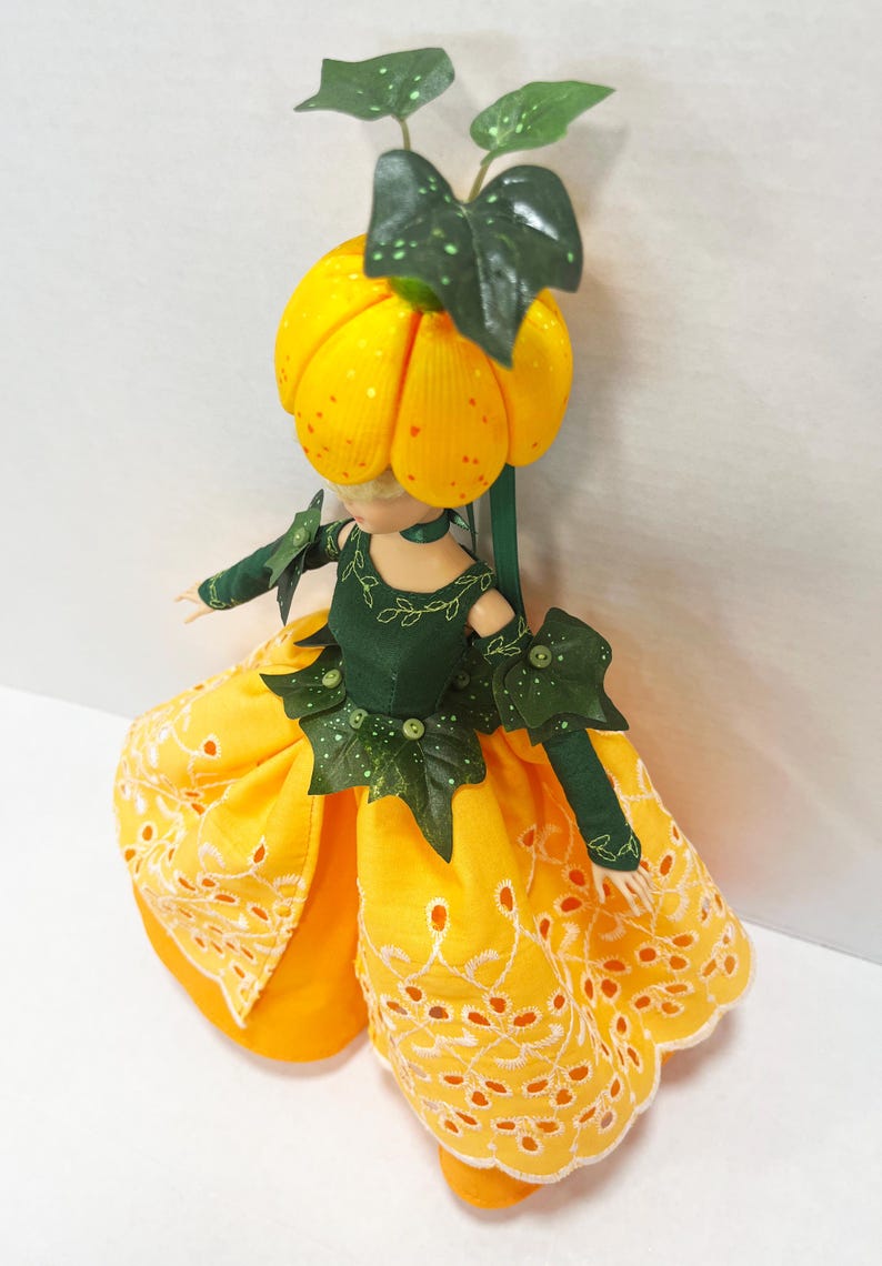 May include: A doll with a yellow pumpkin-shaped head topped with green leaves, wearing a dark green bodice with leaf accents and a bright yellow skirt featuring white eyelet embroidery along the hem, paired with matching green arm sleeves.