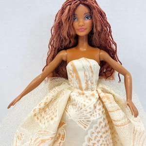 Puede incluir: Mu&ntilde;eca con pelo largo color casta&ntilde;o rojizo y una corona amarilla. La mu&ntilde;eca lleva un vestido sin tirantes con un estampado de conchas en crema y melocot&oacute;n. La falda tiene una capa de tul brillante.