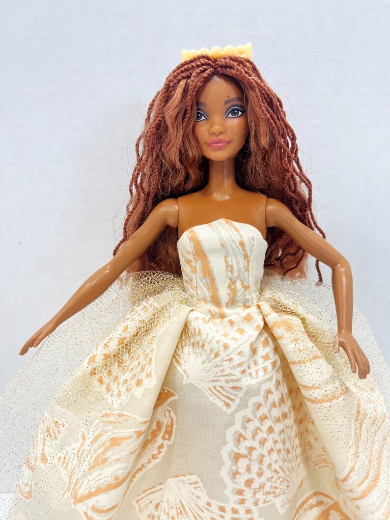 Puede incluir: Mu&ntilde;eca con pelo largo color casta&ntilde;o rojizo y una corona amarilla. La mu&ntilde;eca lleva un vestido sin tirantes con un estampado de conchas en crema y melocot&oacute;n. La falda tiene una capa de tul brillante.