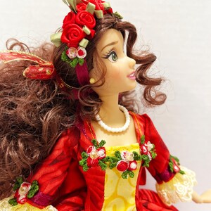 May include: A doll with brown curly hair and green eyes, wearing a red dress with yellow accents and white lace trim. The doll has a pearl necklace and a floral headpiece with red roses and green leaves.