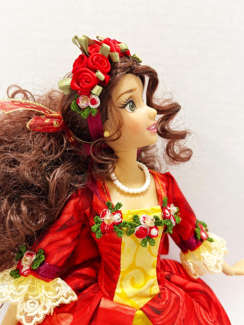 May include: A doll with brown curly hair and green eyes, wearing a red dress with yellow accents and white lace trim. The doll has a pearl necklace and a floral headpiece with red roses and green leaves.
