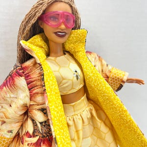 Puede incluir: Mu&ntilde;eca con un atuendo amarillo con dise&ntilde;os de abejas y panales, y una chaqueta con estampado de girasoles. El atuendo incluye un top corto y una falda. La mu&ntilde;eca lleva gafas rosas.