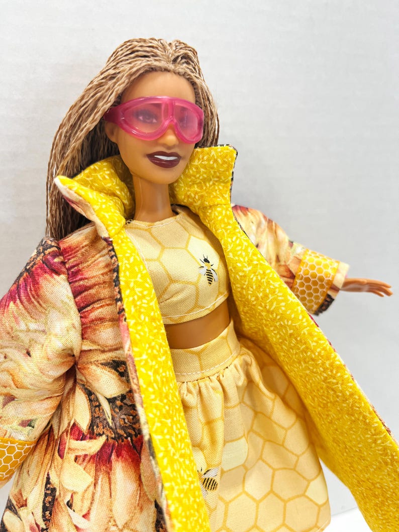 Puede incluir: Mu&ntilde;eca con un atuendo amarillo con dise&ntilde;os de abejas y panales, y una chaqueta con estampado de girasoles. El atuendo incluye un top corto y una falda. La mu&ntilde;eca lleva gafas rosas.