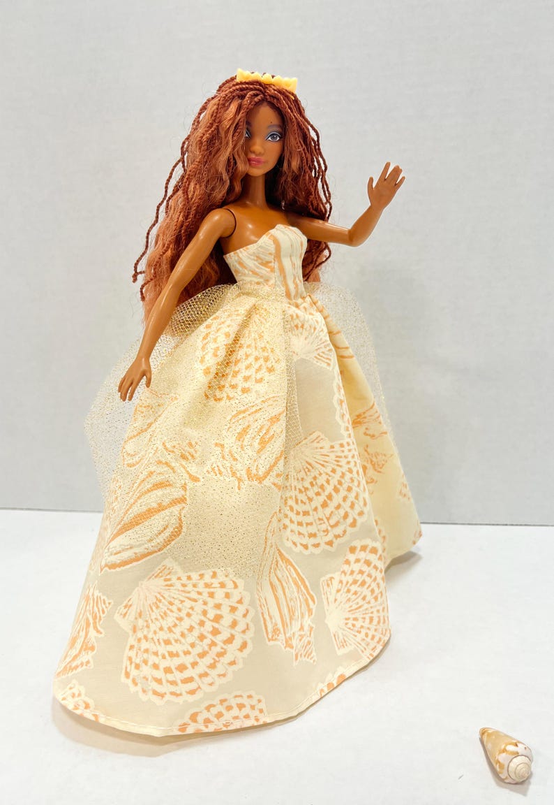 Puede incluir: Mu&ntilde;eca de moda con largo cabello casta&ntilde;o rojizo y una corona amarilla. La mu&ntilde;eca lleva un vestido crema sin tirantes con un estampado de conchas marinas en naranja. El vestido tiene una capa de tul. Una concha est&aacute; en la superficie.