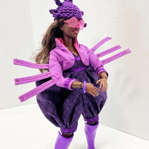 May include: A doll in a purple ensemble, featuring a hat, sunglasses, jacket, dress, and trousers. The hat is adorned with a pink and green crocheted flower. The doll also wears purple boots. The outfit has a unique, artistic design.