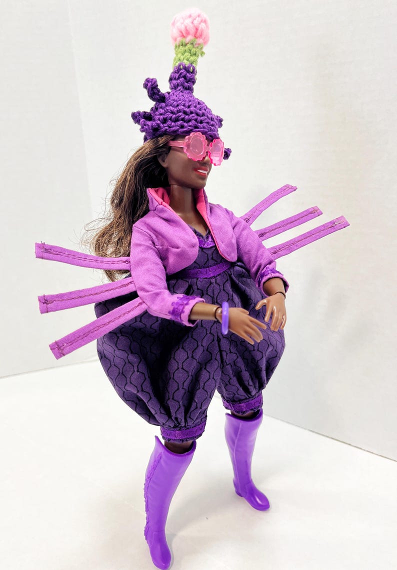 May include: A doll in a purple ensemble, featuring a hat, sunglasses, jacket, dress, and trousers. The hat is adorned with a pink and green crocheted flower. The doll also wears purple boots. The outfit has a unique, artistic design.