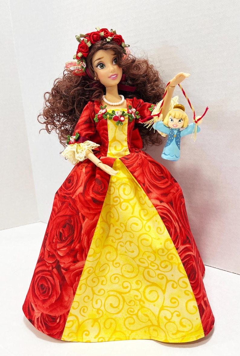 May include: A doll in a red rose-patterned gown with a yellow panel, a floral headpiece, and a small doll. The doll's dress has long sleeves and is trimmed with lace and floral accents. The doll is holding a small doll with a blue dress.