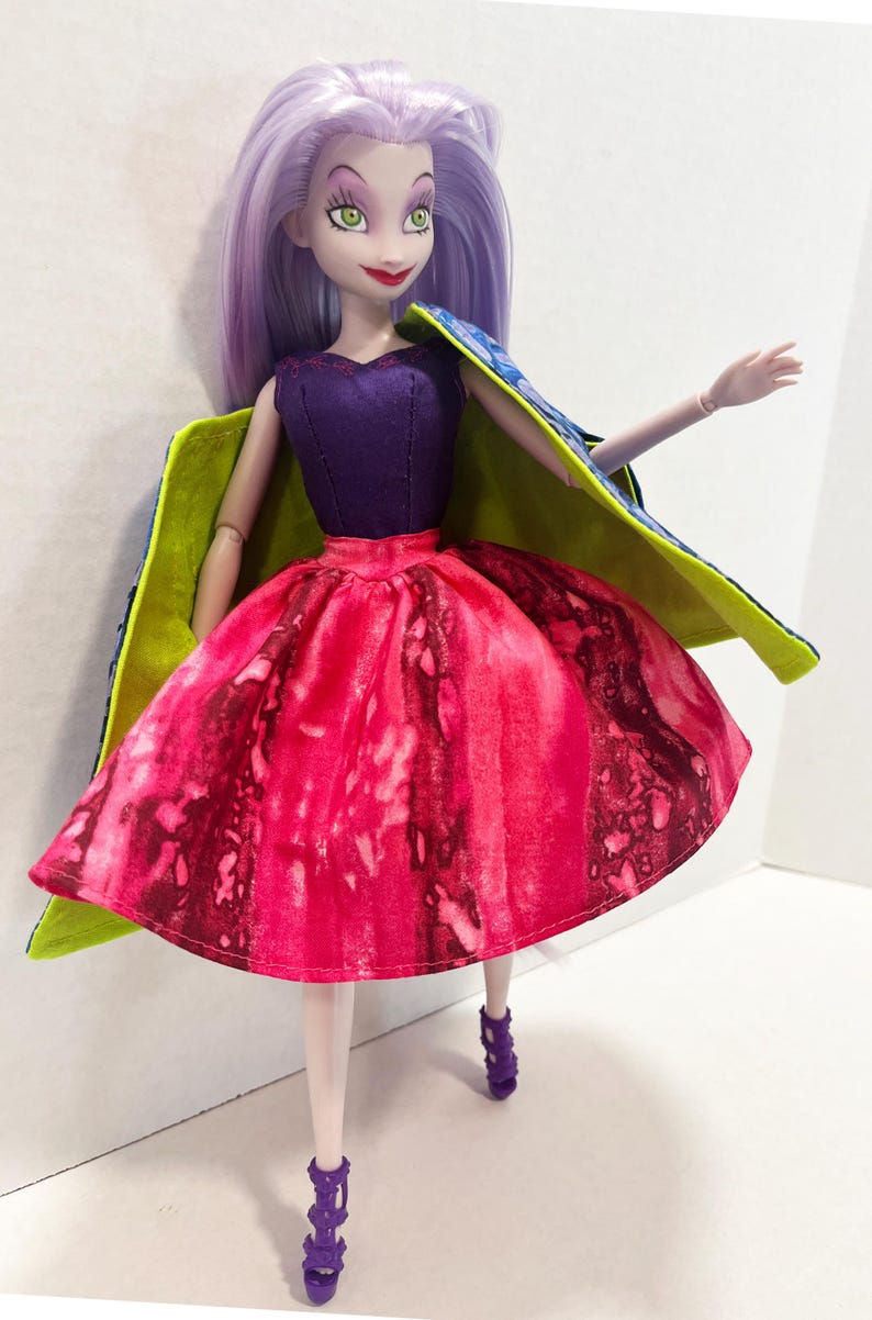 Pu&ograve; includere: Una bambola con lunghi capelli color lavanda, occhi verdi e labbra rosse. La bambola indossa un corpetto senza maniche viola scuro, una gonna con motivo ad acquerello rosa e rosso vivace e un mantello blu con fodera verde lime. Sandali viola.