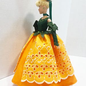 May include: A doll with blonde hair styled in an updo with a green ribbon. The doll wears a dark green bodice with leaf-like accents and long sleeves, paired with a bright yellow skirt featuring a white embroidered eyelet scalloped overlay.