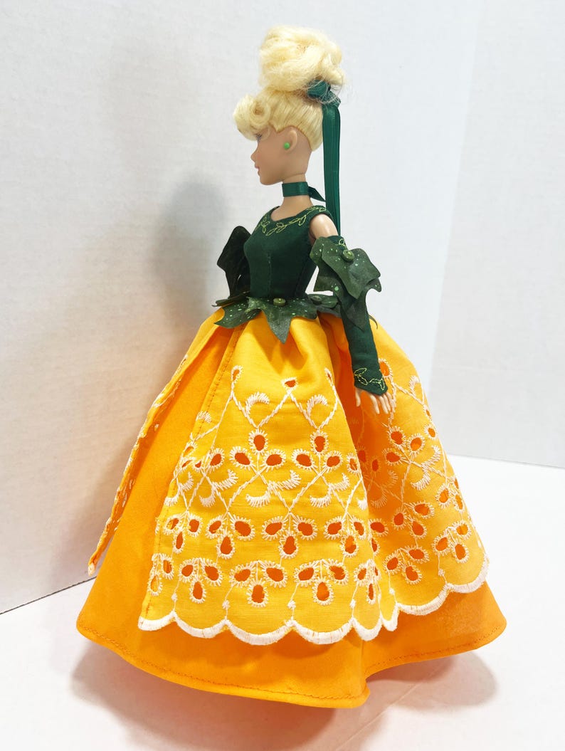 May include: A doll with blonde hair styled in an updo with a green ribbon. The doll wears a dark green bodice with leaf-like accents and long sleeves, paired with a bright yellow skirt featuring a white embroidered eyelet scalloped overlay.