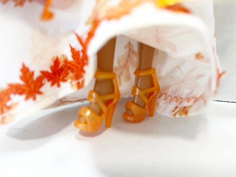 May include: A pair of orange high-heeled sandals with a textured surface and ankle straps, worn by a doll. The doll is wearing a white skirt featuring a repeating pattern of orange and light brown autumn maple leaves along the hemline.