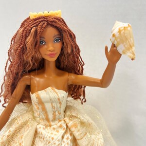 Puede incluir: Mu&ntilde;eca con piel morena, vestida con un vestido crema y naranja sin tirantes y una corona amarilla. La mu&ntilde;eca tiene el pelo largo, casta&ntilde;o rojizo y sostiene una concha. El vestido presenta un estampado de concha.