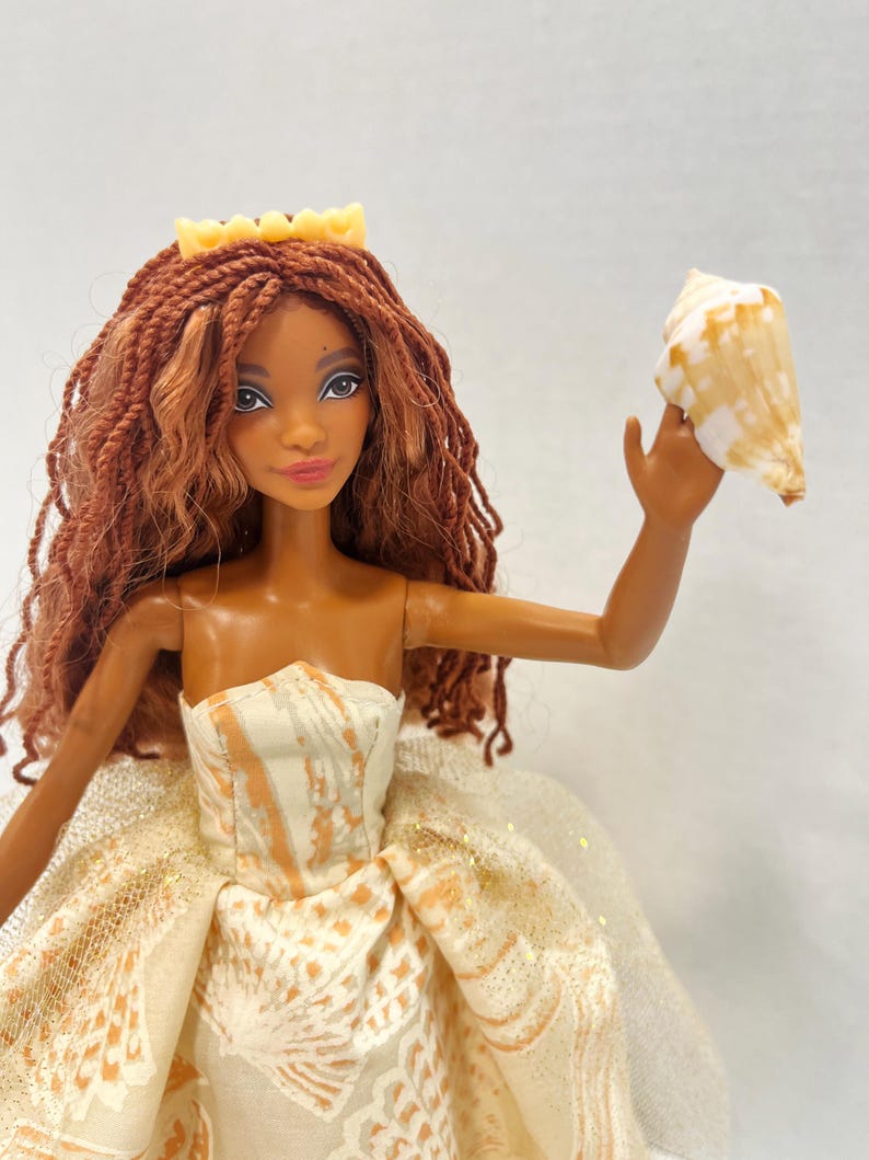 Puede incluir: Mu&ntilde;eca con piel morena, vestida con un vestido crema y naranja sin tirantes y una corona amarilla. La mu&ntilde;eca tiene el pelo largo, casta&ntilde;o rojizo y sostiene una concha. El vestido presenta un estampado de concha.