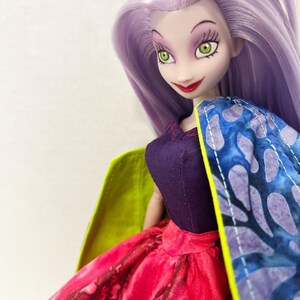 Pu&ograve; includere: Una bambola con lunghi capelli color lavanda, occhi verdi e pelle chiara. La bambola indossa un corpetto viola scuro, una gonna con un vivace motivo rosso e nero e un mantello con stampa blu e viola e una fodera interna verde lime.