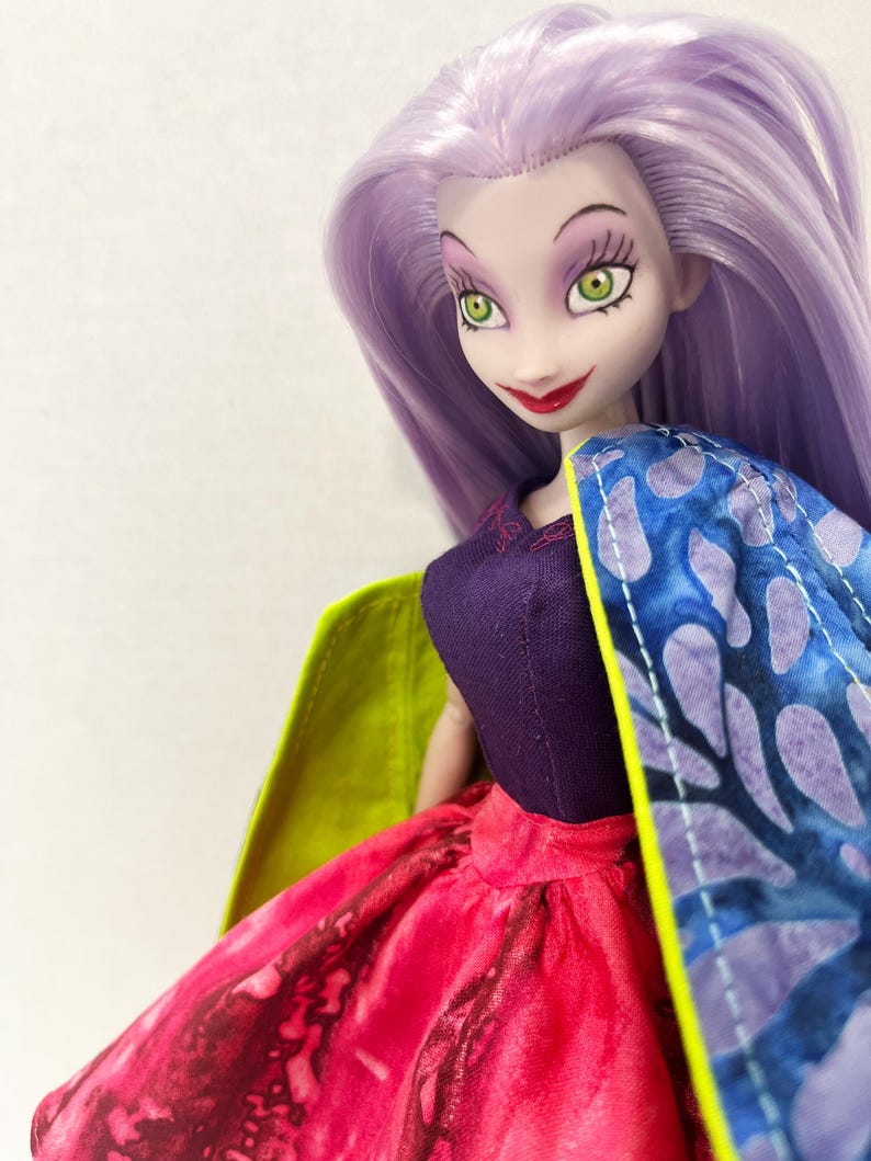 Pu&ograve; includere: Una bambola con lunghi capelli color lavanda, occhi verdi e pelle chiara. La bambola indossa un corpetto viola scuro, una gonna con un vivace motivo rosso e nero e un mantello con stampa blu e viola e una fodera interna verde lime.