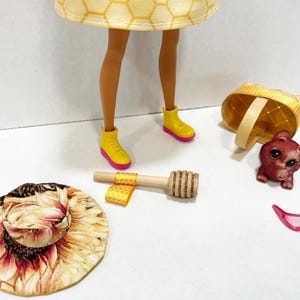 Puede incluir: Una mu&ntilde;eca con un vestido amarillo con estampado de panal y botas amarillas con suelas rosas. Los accesorios incluyen un sombrero de girasol, una cesta peque&ntilde;a, una figura de oso, gafas de sol rosas y un cuchar&oacute;n de miel de madera.