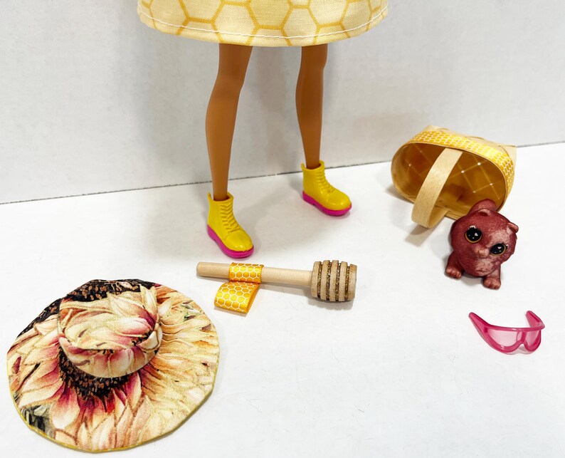 Puede incluir: Una mu&ntilde;eca con un vestido amarillo con estampado de panal y botas amarillas con suelas rosas. Los accesorios incluyen un sombrero de girasol, una cesta peque&ntilde;a, una figura de oso, gafas de sol rosas y un cuchar&oacute;n de miel de madera.
