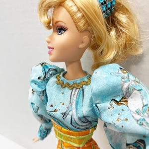 以下が含まれることがあります： ポニーテールに結ばれたブロンドの髪の毛を持つ人形。金色のネックラインとパフスリーブのライトブルーのトップスを着ています。人形はまた、花柄のデザインのオレンジ色のズボンと、宝石付きの青いヘアアクセサリーを身に着けています。