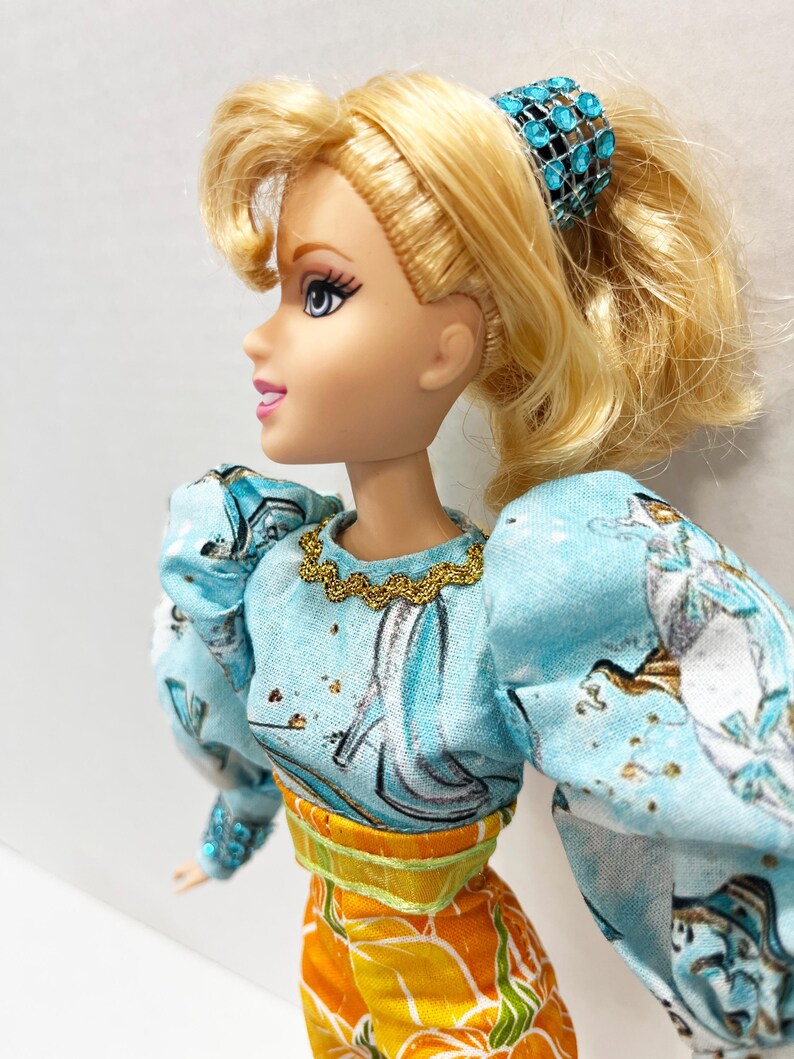 以下が含まれることがあります： ポニーテールに結ばれたブロンドの髪の毛を持つ人形。金色のネックラインとパフスリーブのライトブルーのトップスを着ています。人形はまた、花柄のデザインのオレンジ色のズボンと、宝石付きの青いヘアアクセサリーを身に着けています。