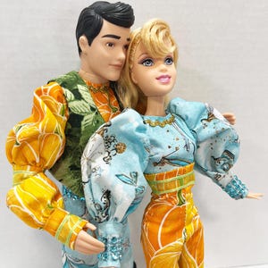以下が含まれることがあります： 男性と女性の人形が、お揃いの衣装を着ています。男性は緑とオレンジの花柄のトップスを、女性は青とオレンジの花柄のトップスとパンツを着用しています。人形は一緒にポーズをとっており、カップルを連想させます。