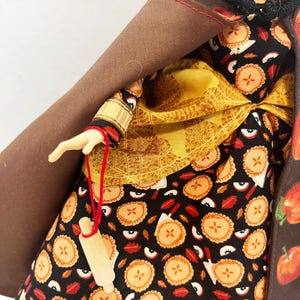 Op de afbeelding: Een pop draagt een zwarte jurk met een patroon van appeltaarten en rode appelschijfjes, een geel gestructureerd schort en een bruine cape met capuchon en appelprint. Een miniatuur houten deegroller hangt aan een rood touwtje aan de hand.