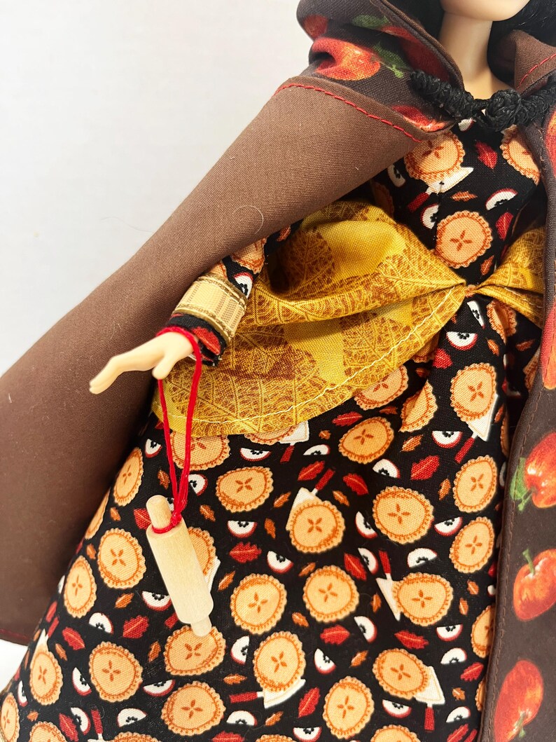Op de afbeelding: Een pop draagt een zwarte jurk met een patroon van appeltaarten en rode appelschijfjes, een geel gestructureerd schort en een bruine cape met capuchon en appelprint. Een miniatuur houten deegroller hangt aan een rood touwtje aan de hand.