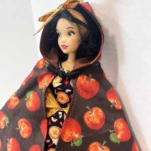 Op de afbeelding: Een pop met zwart haar draagt een bruine cape met capuchon met een rode appelprint en een bijpassende strik. De pop is gekleed in een zwart kledingstuk met een patroon van kleine appeltaartjes. De cape is bij de hals vastgezet met een sluiting.