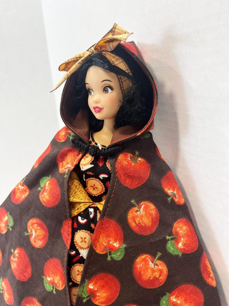 Op de afbeelding: Een pop met zwart haar draagt een bruine cape met capuchon met een rode appelprint en een bijpassende strik. De pop is gekleed in een zwart kledingstuk met een patroon van kleine appeltaartjes. De cape is bij de hals vastgezet met een sluiting.