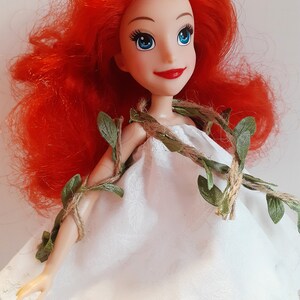 Puede incluir: Una mu&ntilde;eca de pelo rojo con un vestido blanco y una guirnalda de vid verde y cuerda alrededor de los hombros.