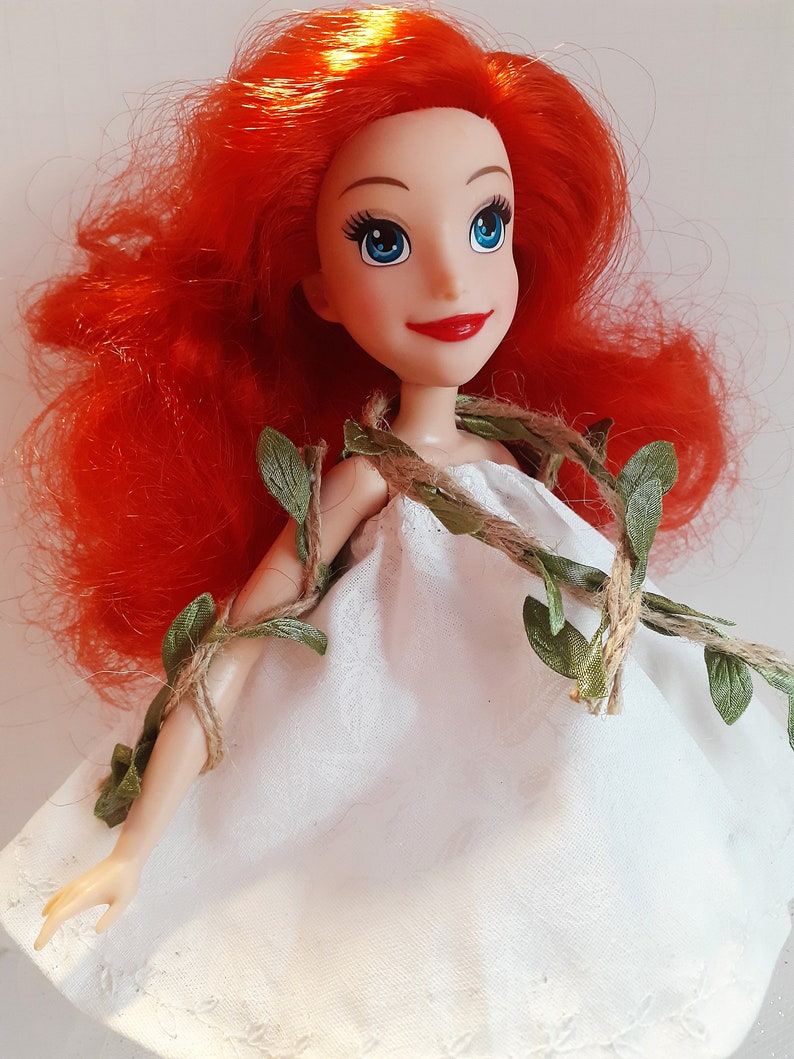 Puede incluir: Una mu&ntilde;eca de pelo rojo con un vestido blanco y una guirnalda de vid verde y cuerda alrededor de los hombros.