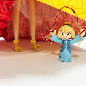May include: A small, hand-painted figurine of a girl in a blue dress with a yellow hair and a gold crown, holding a red and gold rope. The figurine is in front of a red and yellow patterned fabric. Yellow sandals are visible.