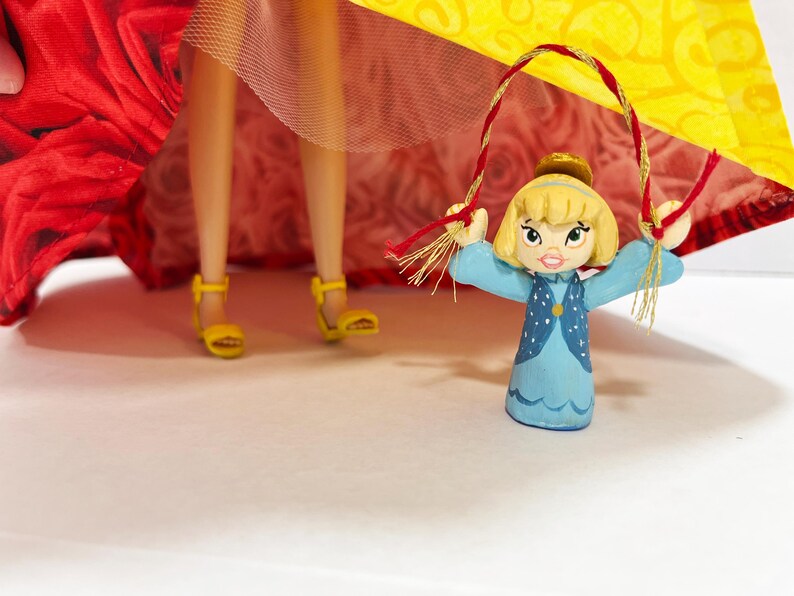 May include: A small, hand-painted figurine of a girl in a blue dress with a yellow hair and a gold crown, holding a red and gold rope. The figurine is in front of a red and yellow patterned fabric. Yellow sandals are visible.