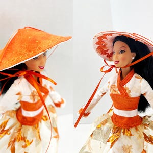 May include: A doll with black hair wearing a white long-sleeved top featuring an orange autumn leaf pattern, a solid orange waistband, and a full skirt with a matching leaf print. The doll wears a wide-brimmed orange hat with a ribbon tie.