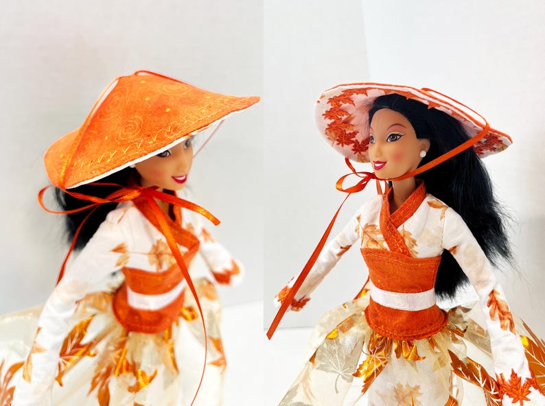 May include: A doll with black hair wearing a white long-sleeved top featuring an orange autumn leaf pattern, a solid orange waistband, and a full skirt with a matching leaf print. The doll wears a wide-brimmed orange hat with a ribbon tie.