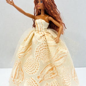 Puede incluir: Mu&ntilde;eca con un vestido sin tirantes color crema con un estampado de conchas marinas en naranja. La mu&ntilde;eca tiene pelo largo color casta&ntilde;o rojizo y una peque&ntilde;a corona dorada. Un velo blanco transparente est&aacute; sujeto al vestido.