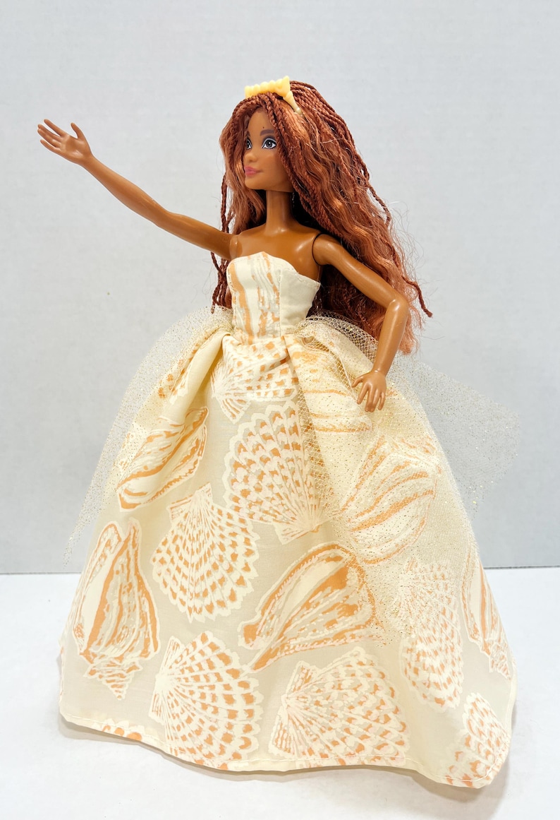 Puede incluir: Mu&ntilde;eca con un vestido sin tirantes color crema con un estampado de conchas marinas en naranja. La mu&ntilde;eca tiene pelo largo color casta&ntilde;o rojizo y una peque&ntilde;a corona dorada. Un velo blanco transparente est&aacute; sujeto al vestido.