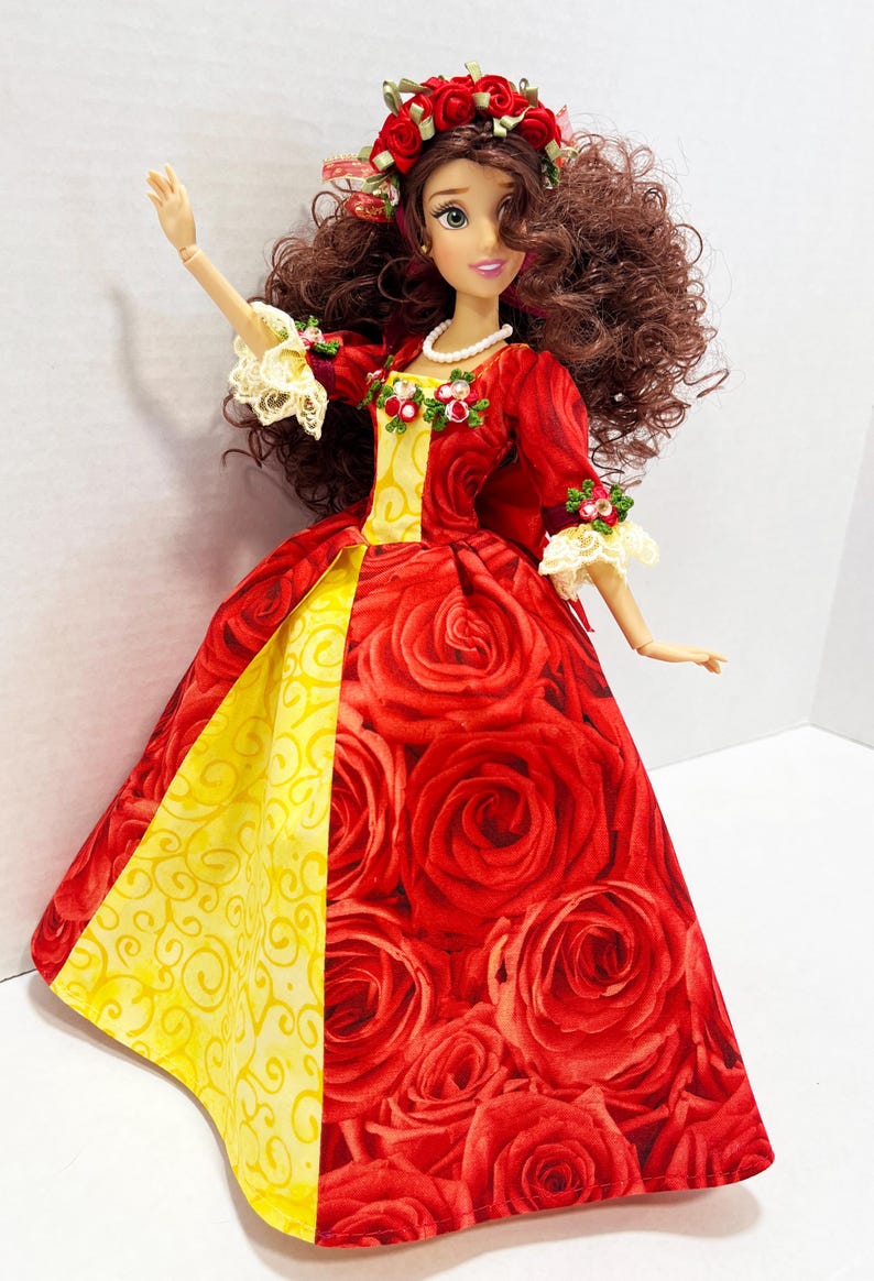 May include: A doll wearing a red gown with a rose pattern, a yellow panel, and a floral crown. The doll has brown curly hair, a pearl necklace, and lace accents on the sleeves. The dress features a full skirt.