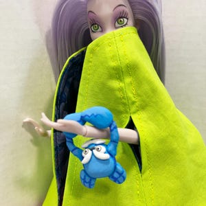 Pu&ograve; includere: Una bambola con lunghi capelli color lavanda e occhi verdi indossa un mantello verde lime brillante con una fodera blu scuro a motivi. La bambola tiene in mano una piccola creatura blu simile a un granchio con dettagli bianchi sul viso.