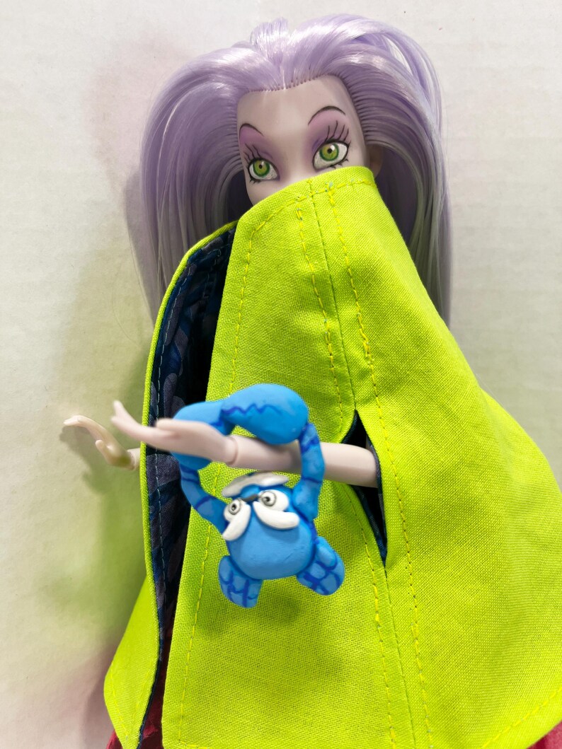 Pu&ograve; includere: Una bambola con lunghi capelli color lavanda e occhi verdi indossa un mantello verde lime brillante con una fodera blu scuro a motivi. La bambola tiene in mano una piccola creatura blu simile a un granchio con dettagli bianchi sul viso.