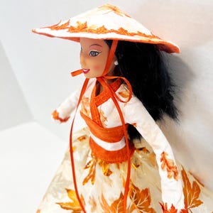 May include: A doll with long black hair wearing a white conical hat and a long-sleeved top featuring orange leaf patterns. The outfit includes a matching full skirt decorated with orange maple leaf prints and orange ribbon ties under the chin.